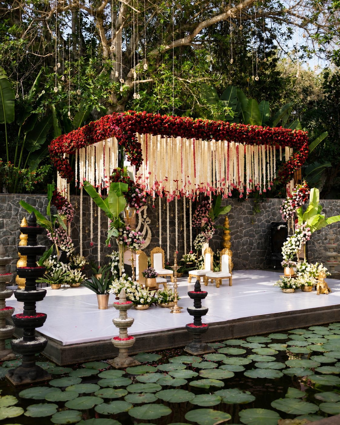 Elegant floral wedding mandap beside a lily pond with red flowers, white garlands, greenery and traditional decor